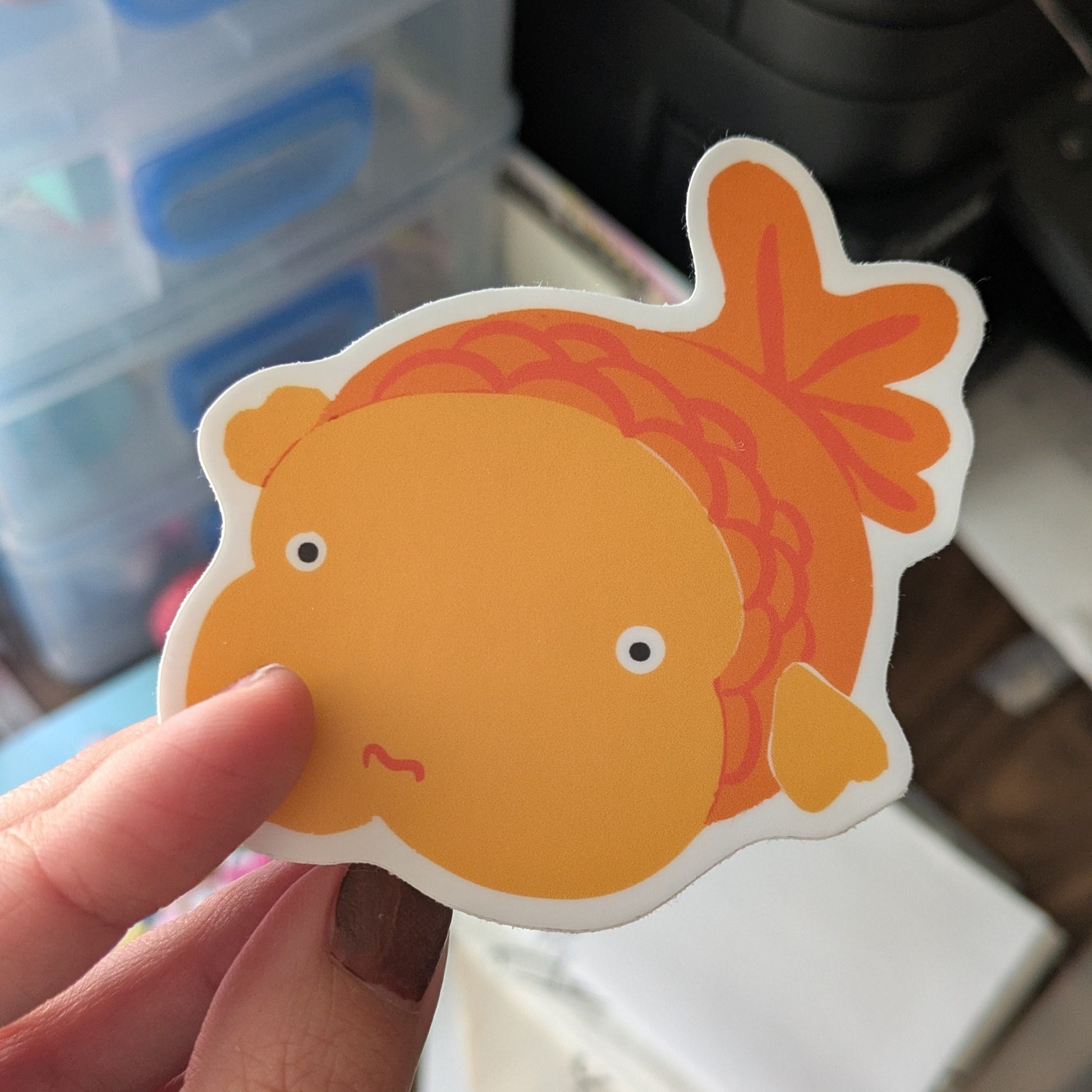 Hand holding a goldfish-shaped sticker with a blurred background