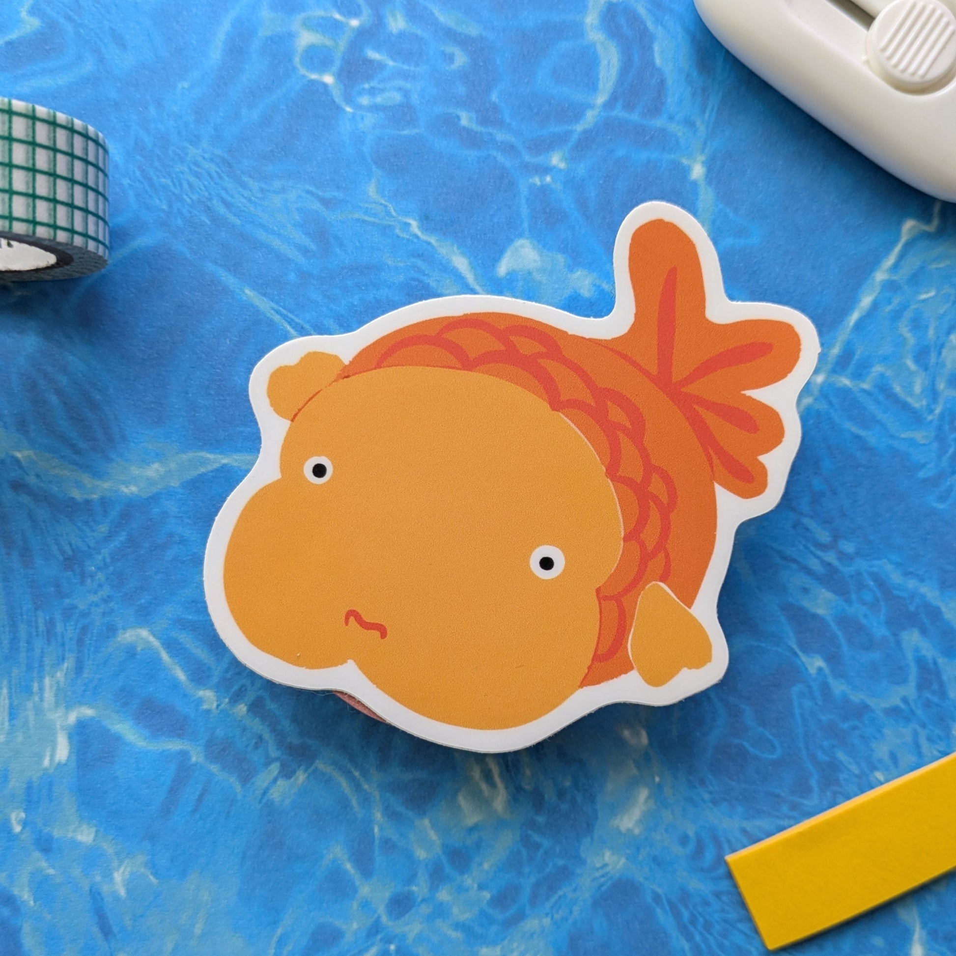 Cartoon goldfish sticker on a blue textured surface with a yellow object and white device.